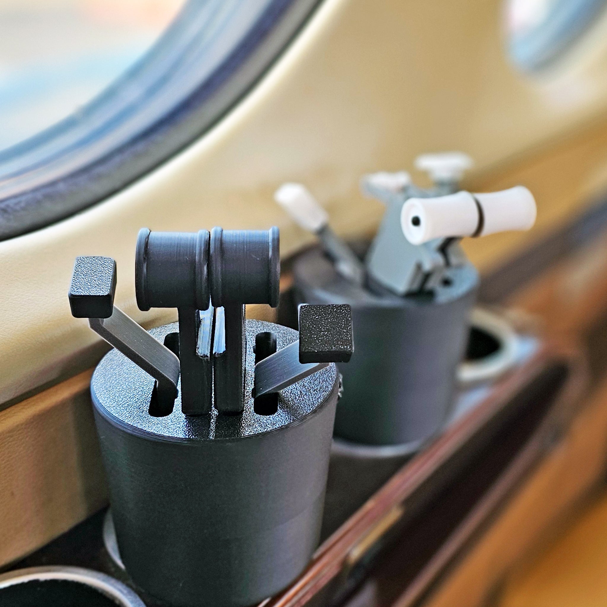Close-up of the Save More! Boeing 737 + Airbus - The Cup Holder Flight Fidget from FlightFidgets.com, showcasing the throttle controls inside a cockpit near a window. The levers and knobs are positioned in holders, providing airplane enthusiasts with a tactile experience, as the sky can be seen through the window. The equipment features metallic construction with some plastic components.