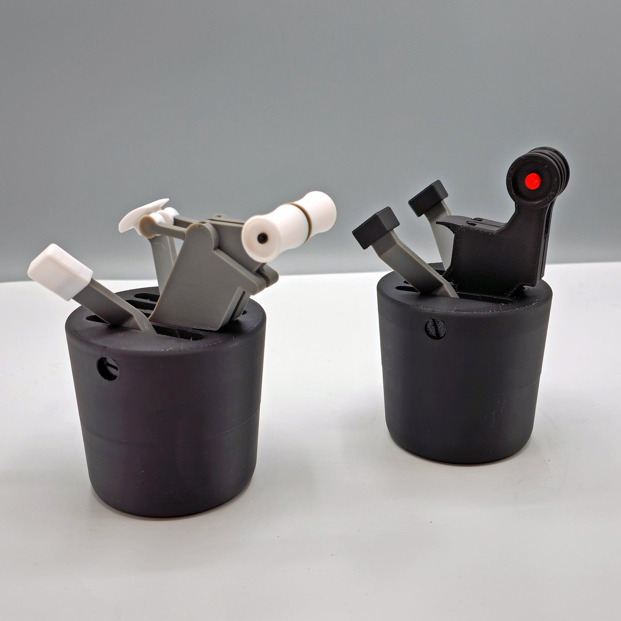 The Save More! Boeing 737 + Airbus - The Cup Holder Flight Fidget by FlightFidgets.com presents a captivating sensory experience with two black cylindrical designs featuring mechanical levers and components against a gray backdrop. The piece on the left incorporates white and gray elements, while the one on the right showcases a bold black and red color palette—ideal for airplane enthusiasts seeking distinctive design appeal.