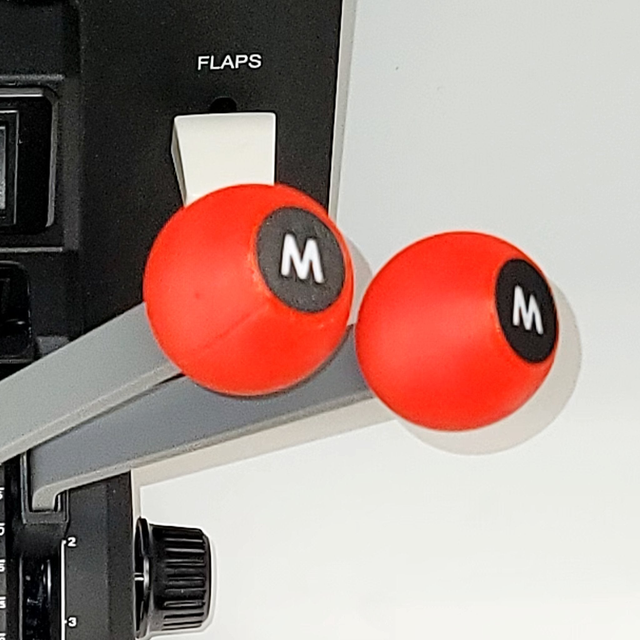 A close-up of the Prodesksim DC-3 set of 6 Levers Addon for the Honeycomb Bravo Throttle Quadrant highlights red and gray throttle levers with round handles marked "M." In the background, a switch labeled "FLAPS" is visible. Reminiscent of a classic DC3 Twin Engine, this panel contrasts with the colorful levers, including 2X throttle, 2X mixture, and 2X prop controls.