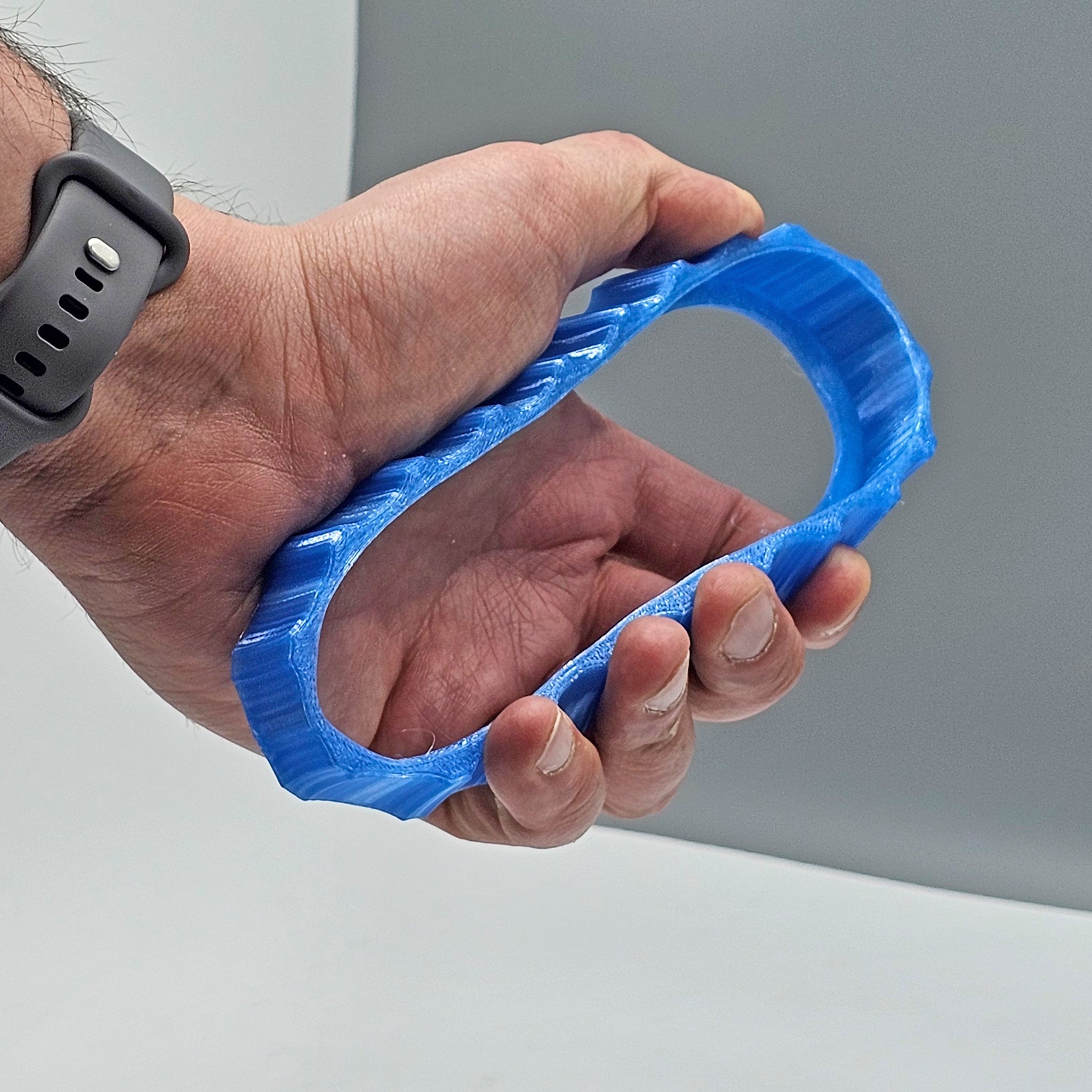 In a plain grey background, a hand displays a vibrant blue 3D-printed object crafted by Prodesksim: the 1 Color Vibrant and Flexible Trim Wheel Cover, designed for the Honeycomb Bravo Throttle Quadrant. The cover, with its oval shape and textured edges, stands out against the wrist of the person wearing a black smartwatch, likely customized for flight simulation.