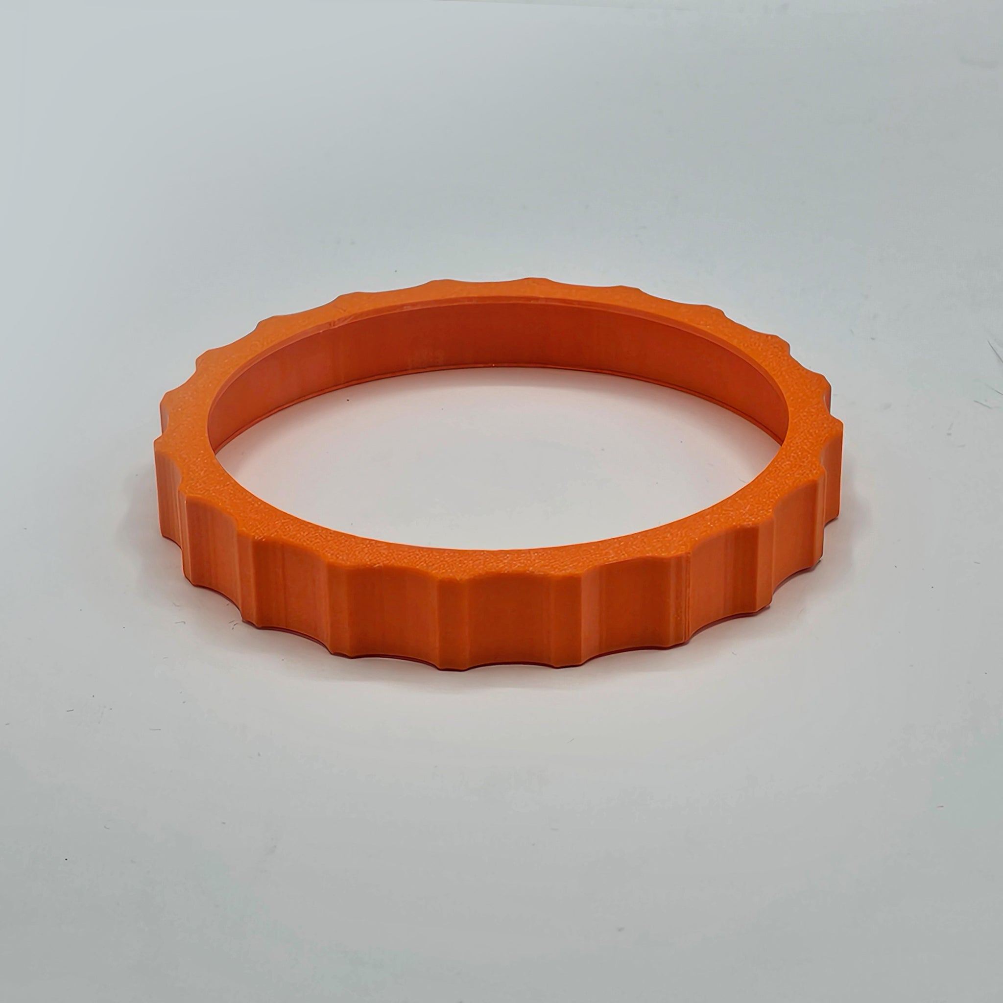 A vibrant orange, textured plastic ring, designed to resemble a gear, sits on a smooth light gray surface. This "1 Color Vibrant and Flexible Trim Wheel Cover" by Prodesksim features evenly spaced ridges around its outer edge, making it perfect for enhancing the tactile experience of your Honeycomb Bravo Throttle Quadrant during flight simulation.