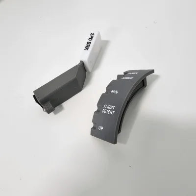 Two gray control levers by Prodesksim rest on a sleek white surface: one with a curved Boeing 737 design marked "FLIGHT DETENT" at 50% and UP, and the other, part of their Boeing 737/757/767/777/787 series, is a spring-loaded Speed Brake labeled "SPD BRK" with a white tip.