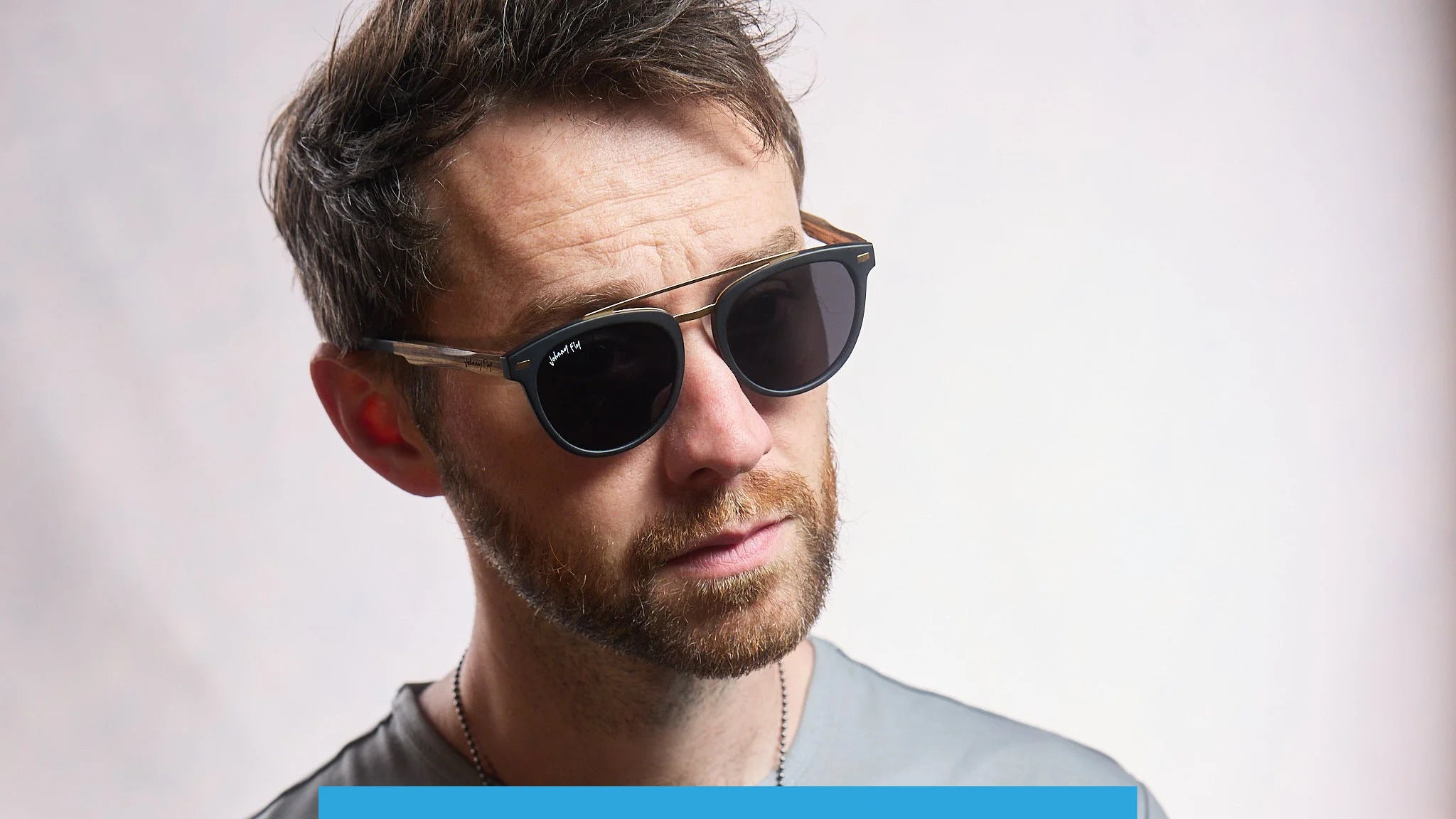 The man with short hair and a beard is wearing the Johnny Fly Captain sunglasses, which boast a sleek acetate front and wooden arms. His neutral expression is highlighted by the gently polarized lenses, all set against a plain, softly lit background as he gazes slightly to the side.