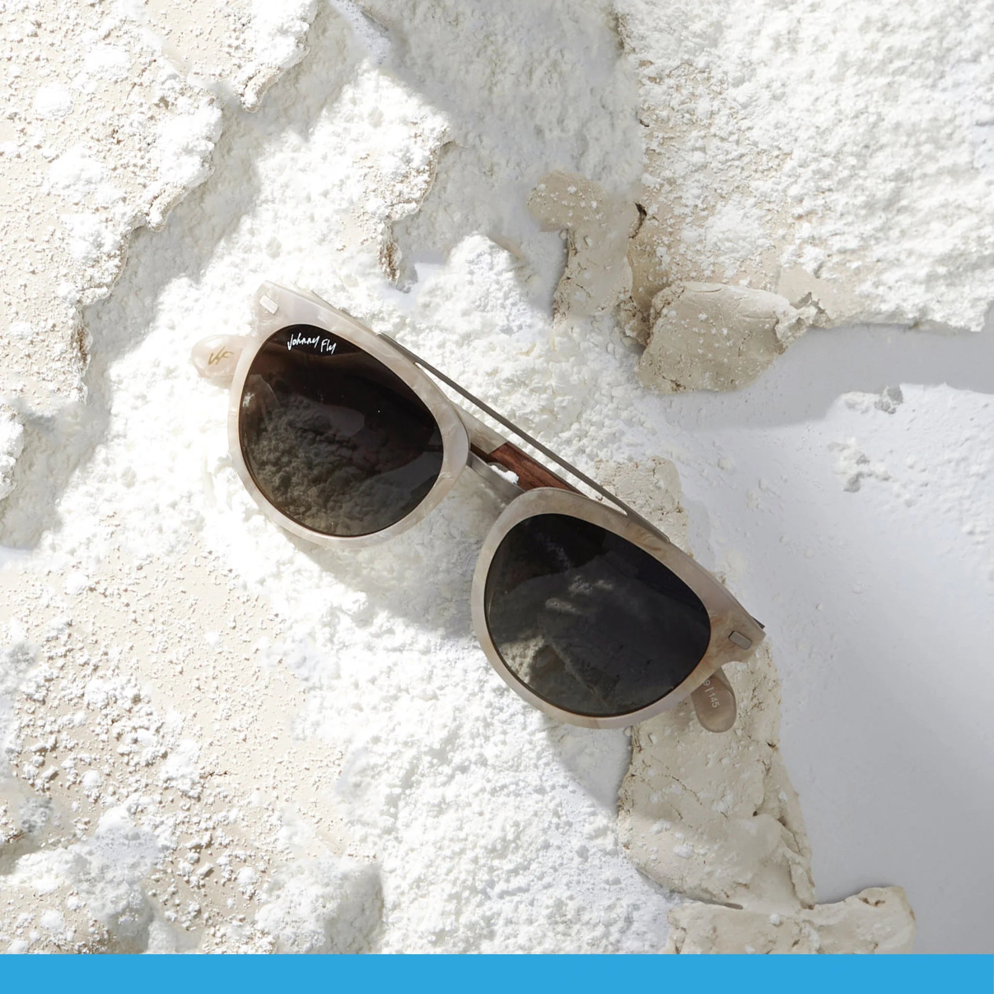 A pair of Johnny Fly's Captain sunglasses, featuring beige frames and dark polarized lenses, rests on a textured surface dusted with white powder. Shadows cast across the scene emphasize the modern design, accentuated by sleek wooden arms.