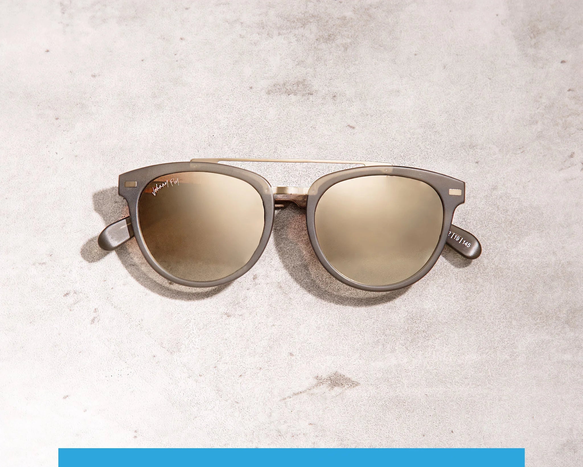 The Captain sunglasses from Johnny Fly, featuring polarized lenses, an acetate front section, and a metallic frame, rest on a light gray textured surface. The earpieces are sleek and modern.