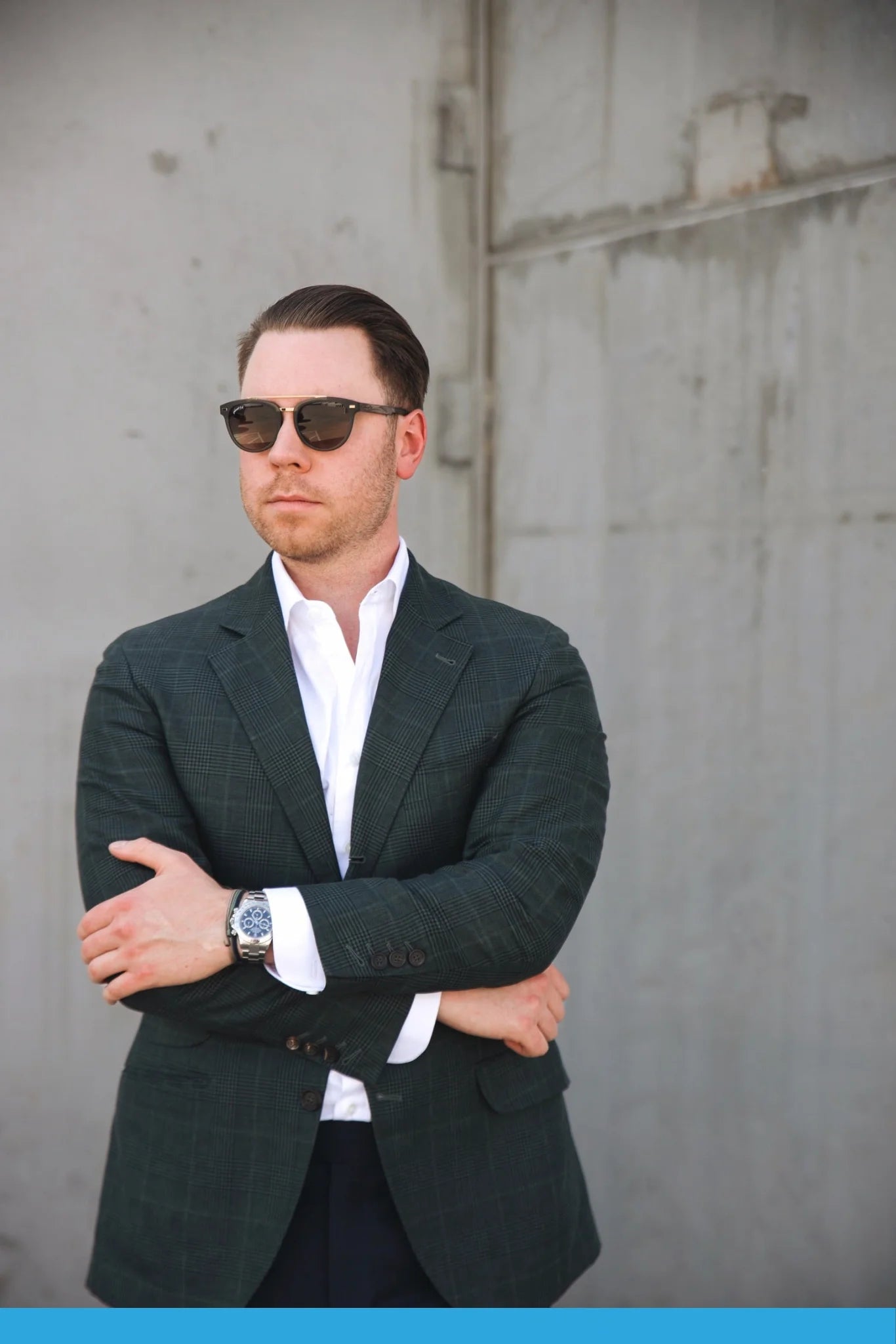 A person confidently standing in front of a concrete wall sports the Johnny Fly Captain sunglasses, featuring an acetate front section and wooden arms, paired with a dark green blazer and a white shirt with folded arms.