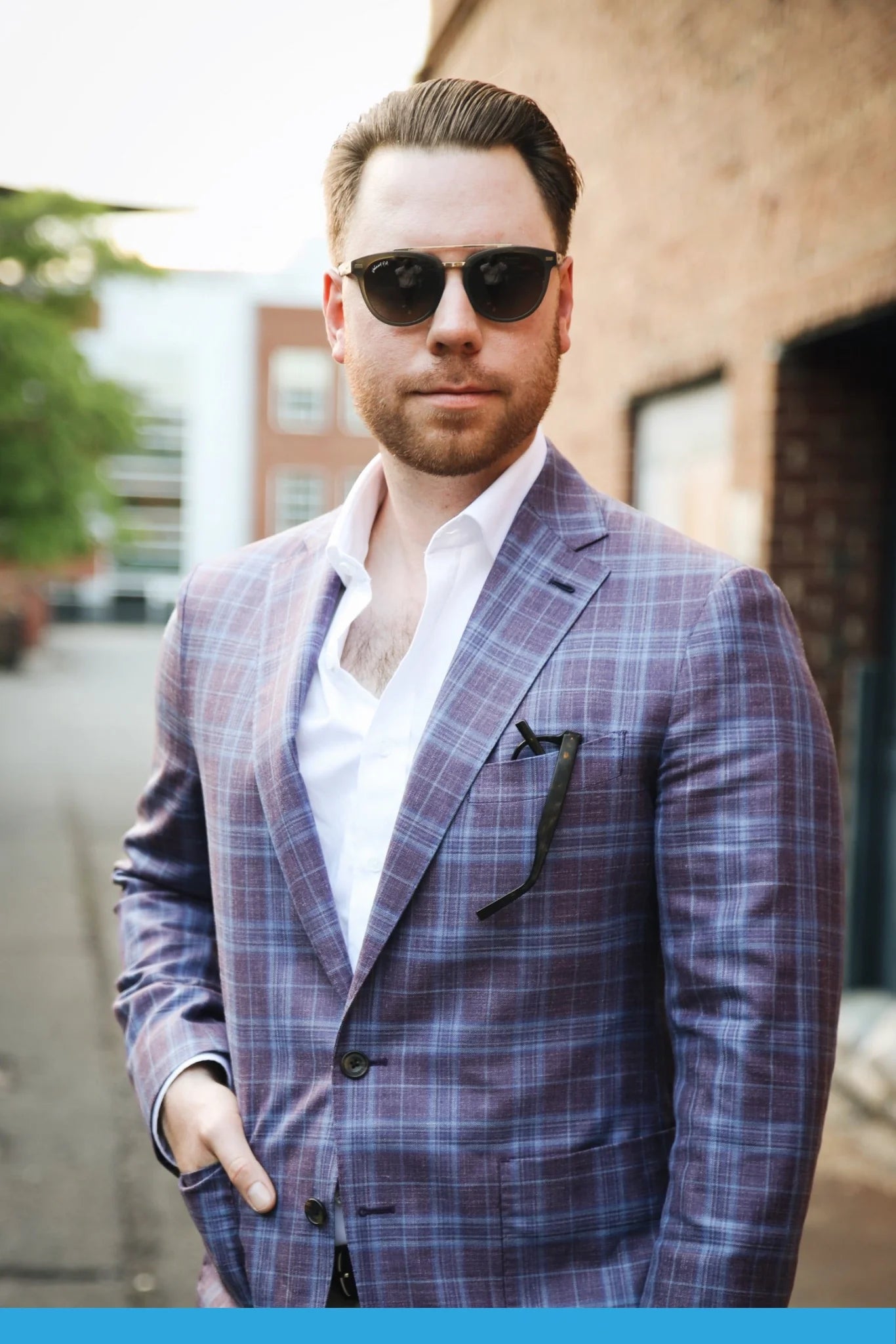 A man exudes confidence in a purple plaid suit, with one hand in his pocket and wearing the "Captain" sunglasses by Johnny Fly. These sunglasses feature an acetate front section, wooden arms, and polarized lenses that glint subtly against the urban backdrop of brick buildings and lush greenery.