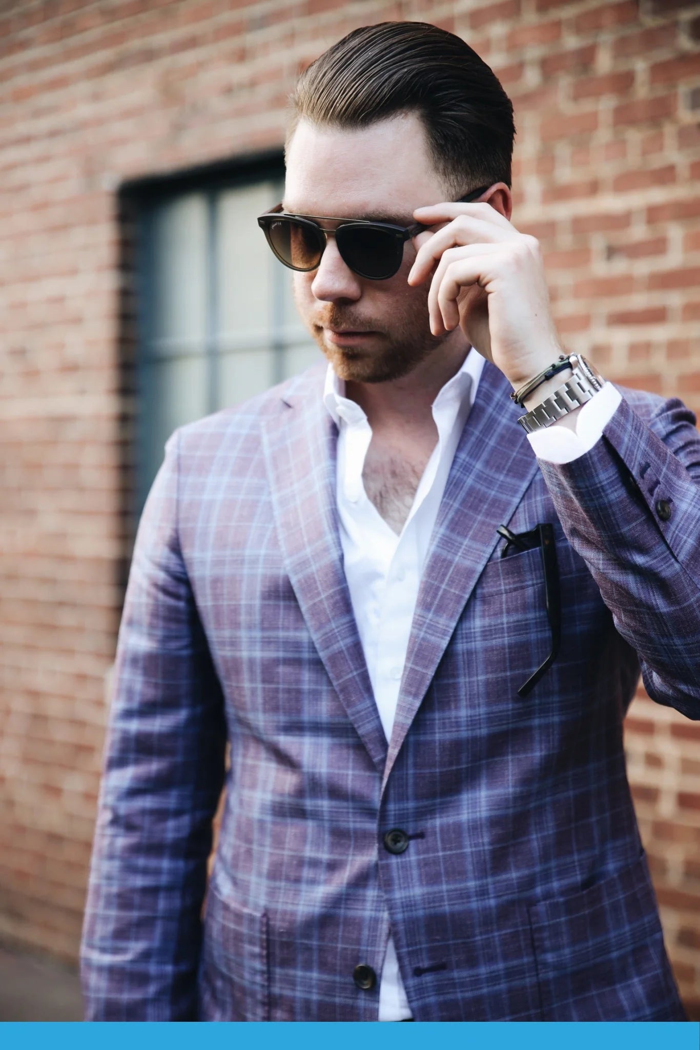 A person wearing the Captain sunglasses by Johnny Fly, featuring polarized lenses and wooden arms, adjusts their eyewear. They stand in front of a brick wall, with a purple plaid suit and a white shirt partially visible underneath the acetate front section of the suit jacket.