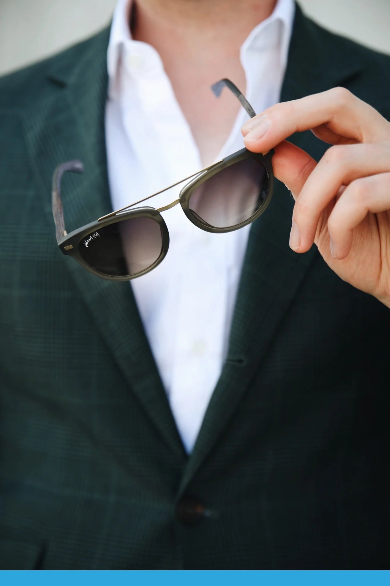 A person dressed in a dark green plaid suit and white shirt holds up a pair of Johnny Fly's Captain sunglasses, highlighting the polarized lensing and stylish acetate front section. The blurred background enhances the focus on the suit and eyewear.