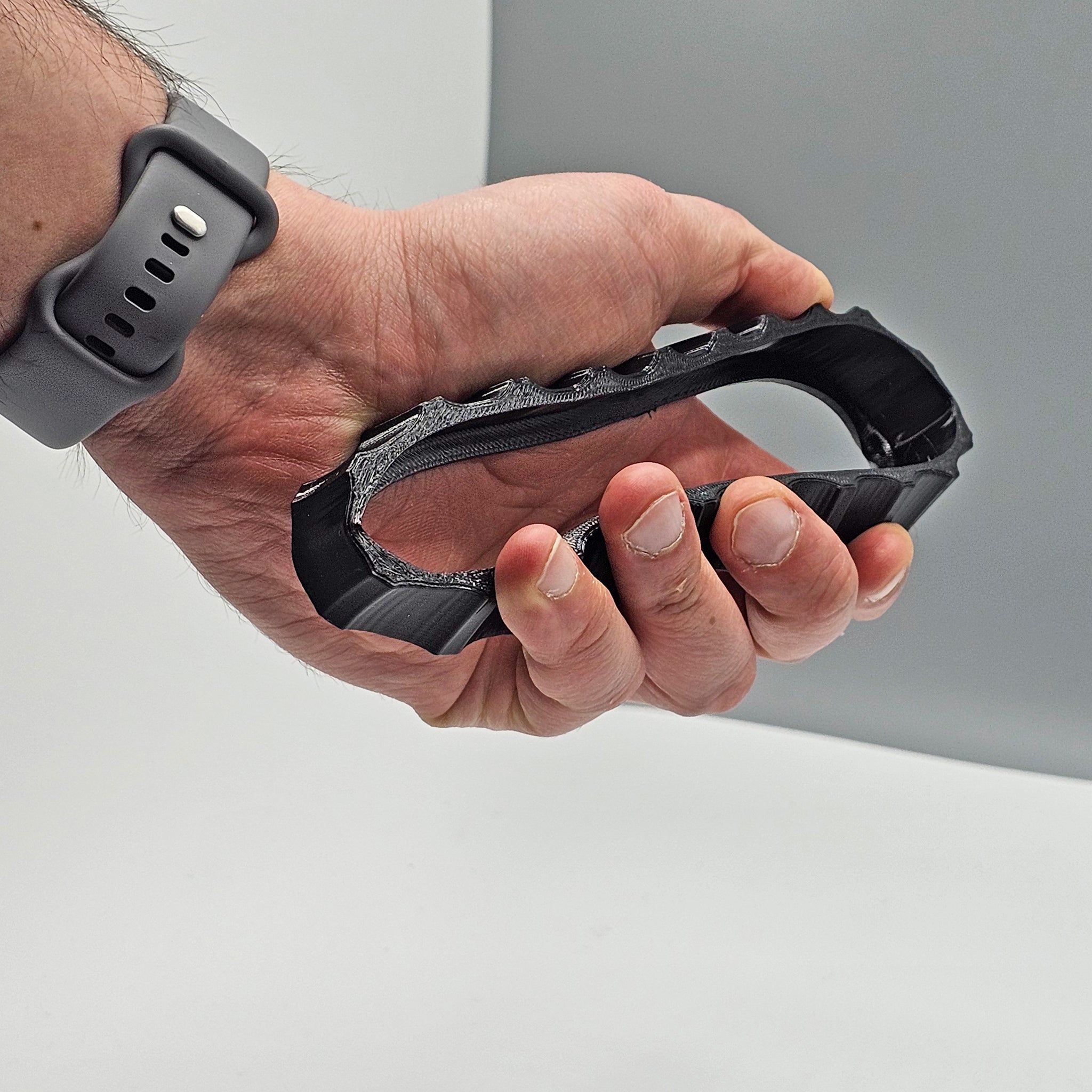 A person wearing a gray watch demonstrates holding a black, textured grip similar to the ergonomic design found in the Prodesksim 1 Color Vibrant and Flexible Trim Wheel Cover for Honeycomb Bravo Throttle Quadrant. The object is held with fingers inside, set against a plain background.