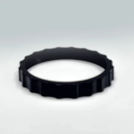 On a smooth, light gray surface sits a black, circular gear-like object adorned with evenly spaced notches, reminiscent of the 1 Color Vibrant and Flexible Trim Wheel Cover for the Honeycomb Bravo Throttle Quadrant by Prodesksim. Its subtle shadow gently adds depth, invoking images of precision in flight simulation.
