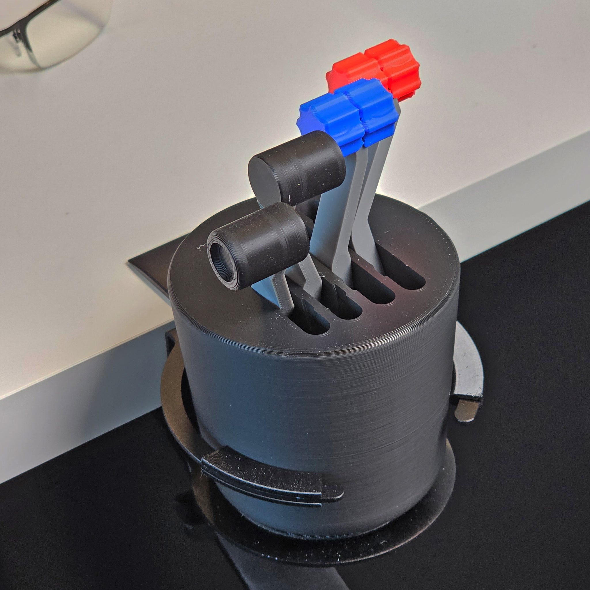 Introducing the "G58 Baron - The Cup Holder Flight Fidget" by FlightFidgets.com: a uniquely designed 3D-printed black cylindrical holder tailored for airplane enthusiasts. This innovative gadget features three joystick-like levers adorned with colorful handles—two in blue and one in red. Perfectly mounted on a flat surface, it serves both as an intriguing fidgeting tool and a flight-themed cup holder.