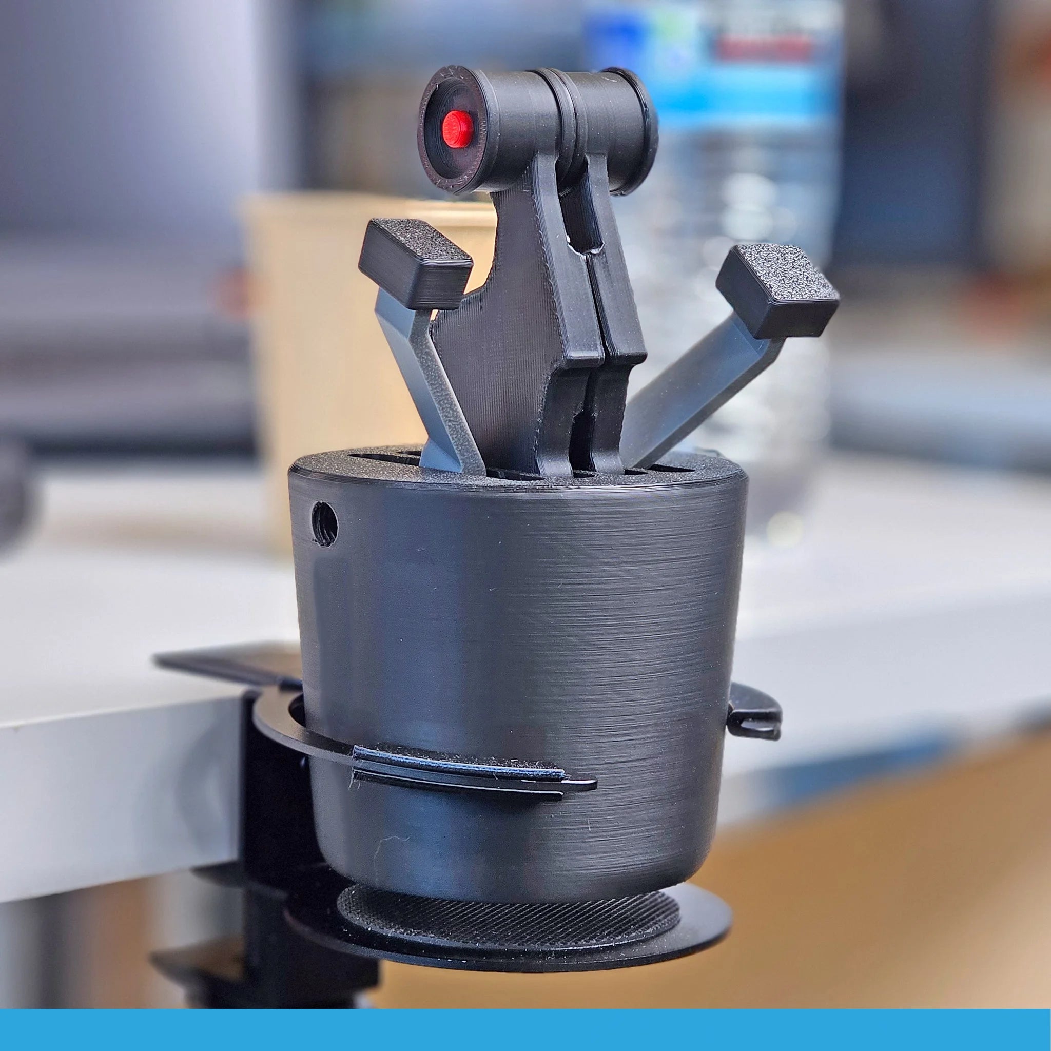 A black 3D-printed AirBus 320 - The Cup Holder Flight Fidget from FlightFidgets.com is clamped to the edge of a desk, ideal for airplane enthusiasts who enjoy manipulating throttle levers. It features three jointed fingers, one with a distinctive red accent. In the background, a water bottle and paper cup are slightly out of focus.
