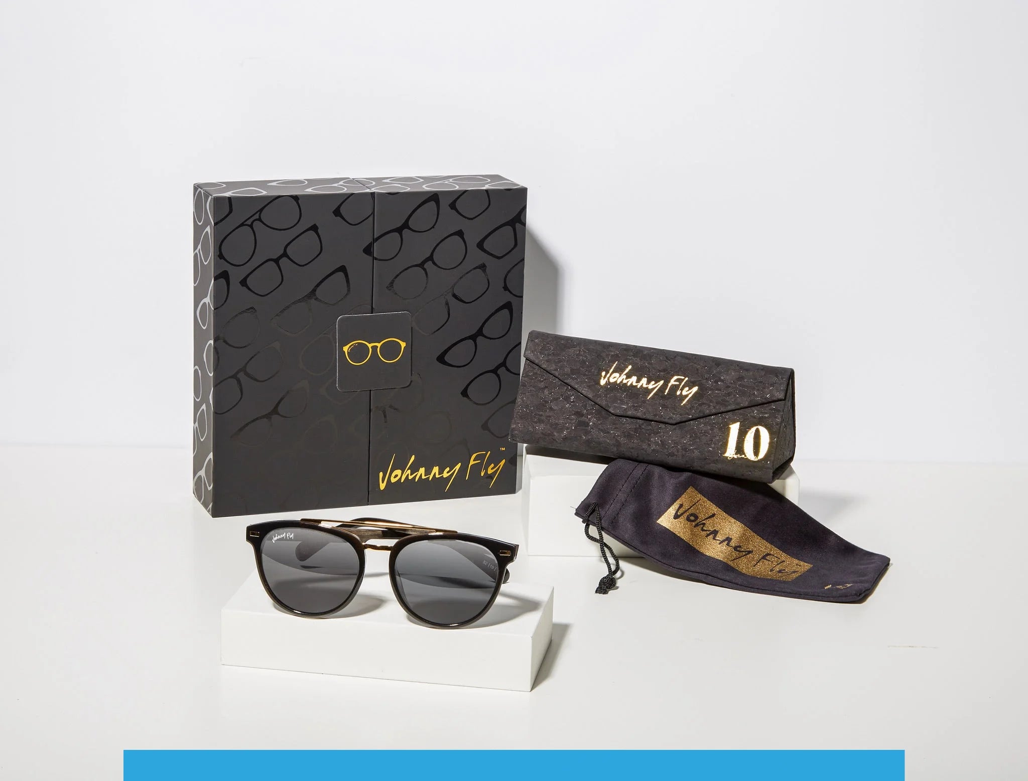 The "Captain" sunglasses by Johnny Fly are showcased with a stylish black and gold-patterned box, a black triangular eyeglass case bearing the "Johnny Fly" logo, and a soft pouch. They feature dark polarized lenses and a tortoiseshell frame complemented by wooden arms, all elegantly presented on a white stand.