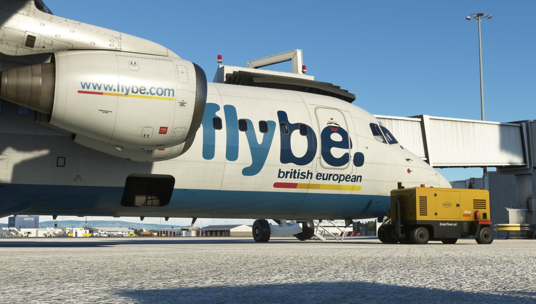 A close-up of a Flybe aircraft parked at an airport gate features "flybe.com" on its engine and "flybe British European" on the fuselage. A ground power unit is stationed nearby, set against a clear blue sky—an image evocative of Just Flight's BAE-146 Professional for MSFS2020.