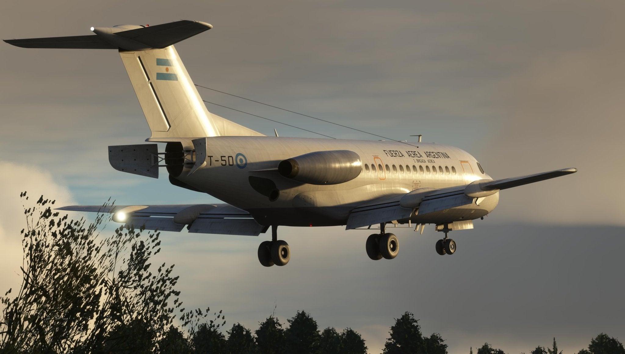 A military aircraft, reminiscent of the Just Flight Fokker-28 Professional for MSFS2020 by Just Flight, has its landing gear extended as it flies low over the trees. The plane's white and gray exterior with blue markings stands out against a cloudy sunset sky.