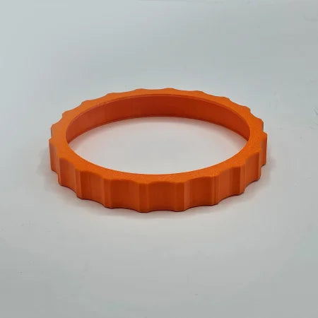 A vibrant orange, textured plastic ring, designed to resemble a gear, sits on a smooth light gray surface. This "1 Color Vibrant and Flexible Trim Wheel Cover" by Prodesksim features evenly spaced ridges around its outer edge, making it perfect for enhancing the tactile experience of your Honeycomb Bravo Throttle Quadrant during flight simulation.