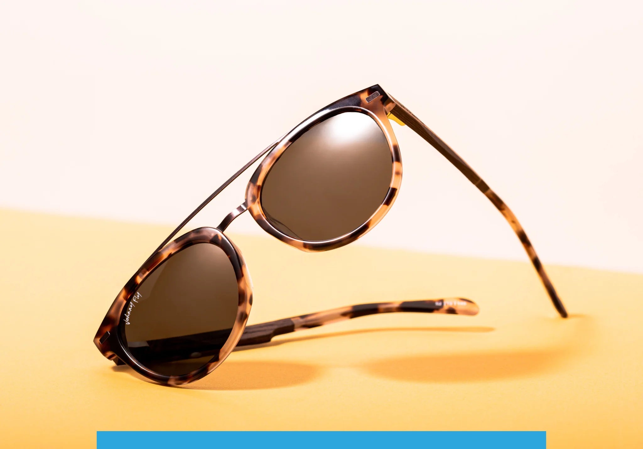 The Captain sunglasses by Johnny Fly, featuring a tortoiseshell design with round frames and an acetate front section, are elegantly positioned on a flat yellow surface. The dark, polarized lenses and wooden arms are tilted to highlight their stylish shadows against the simple backdrop.