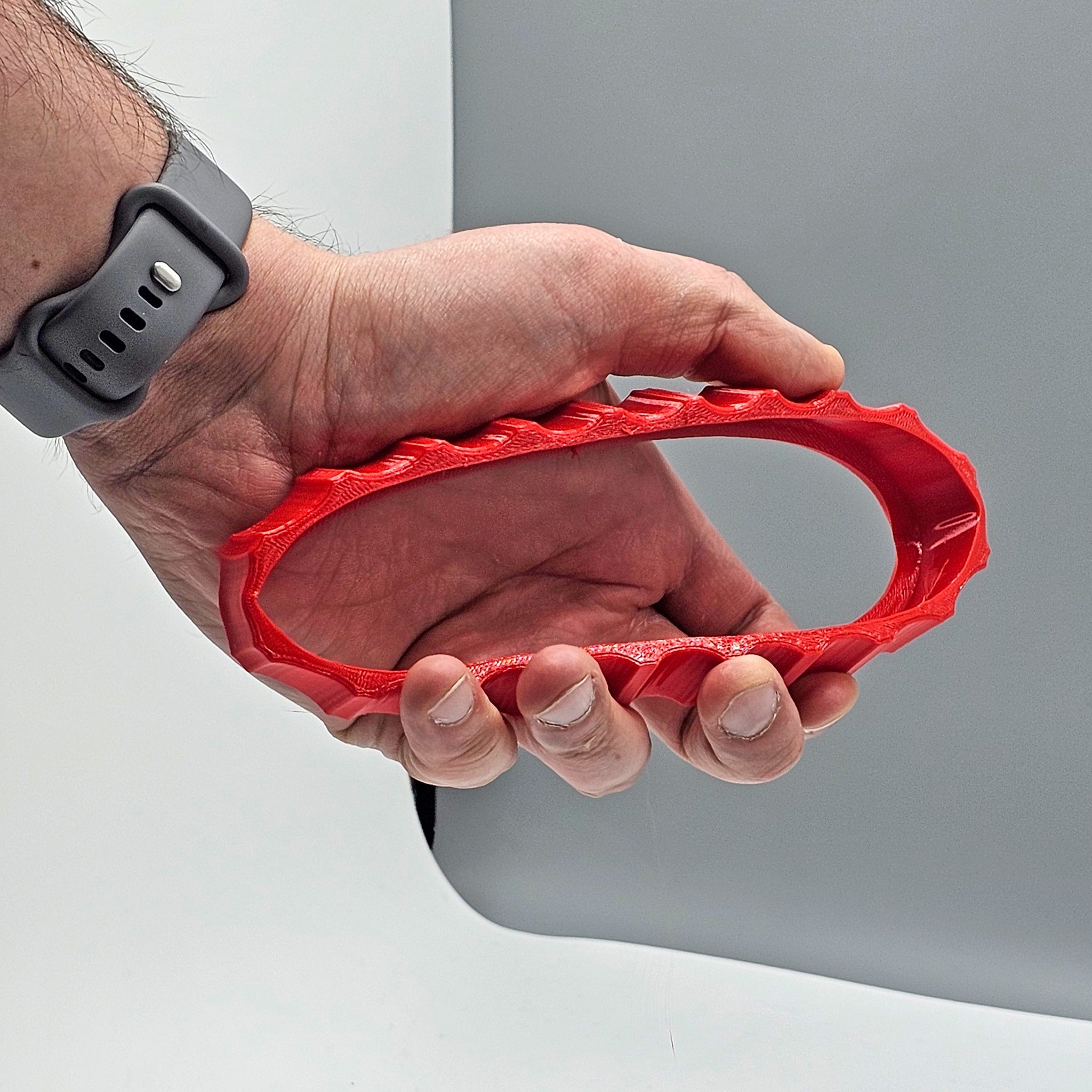 A hand holding the Prodesksim 1 Color Vibrant and Flexible Trim Wheel Cover, featuring a red oval-shaped design with textured edges—perfect for enhancing your Honeycomb Bravo Throttle Quadrant. The muted gray surface in the background complements its sleek design.