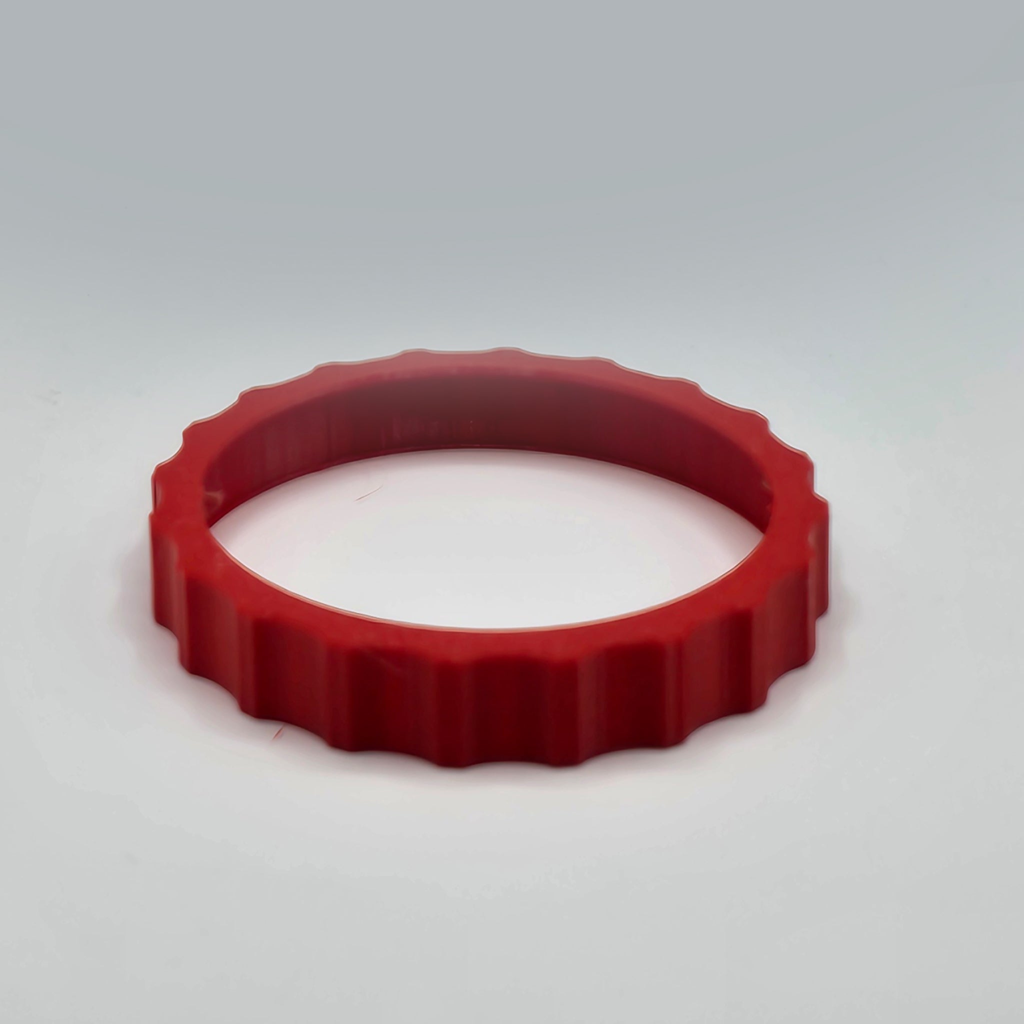 A vibrant red, round plastic object with serrated edges, resembling a bottle cap, is set against a plain gray background. It appears to be the 1 Color Vibrant and Flexible Trim Wheel Cover designed for the Honeycomb Bravo Throttle Quadrant by Prodesksim.