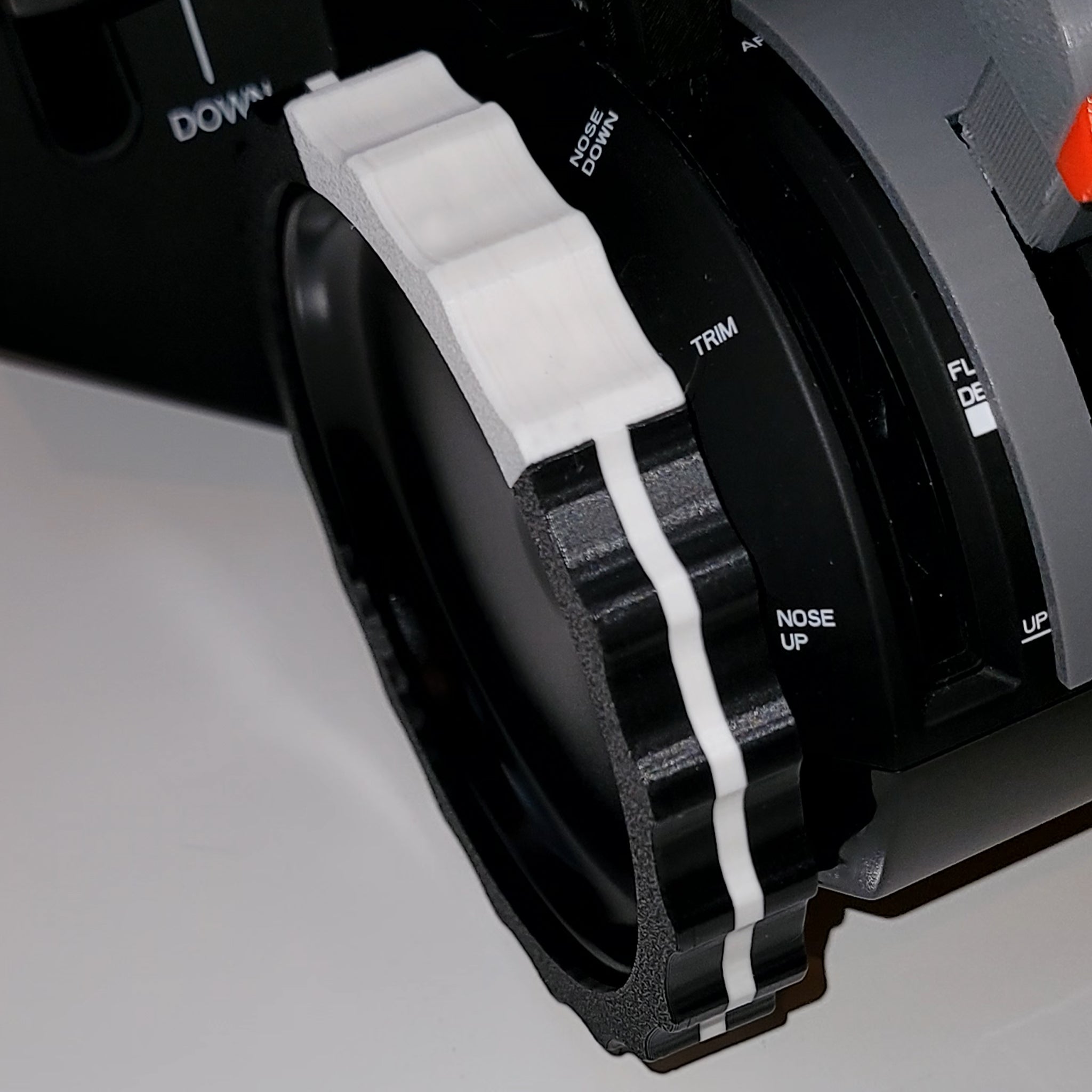 A close-up of a camera lens highlights a black and white striped grip, reminiscent of the Prodesksim's 2 Color TRIMER, a vibrant trim wheel cover for the Honeycomb Bravo Throttle Quadrant. The lens features controls labeled "TRIM" and "NOSE UP," indicating it's designed for advanced flight simulation equipment.