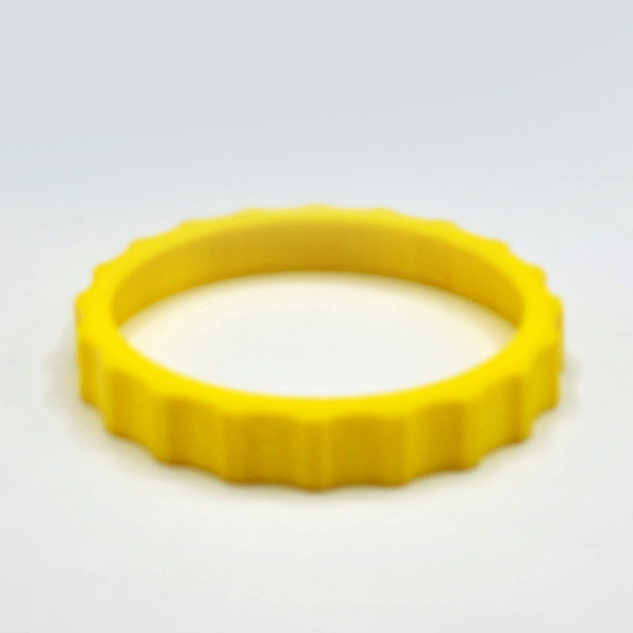 A vibrant yellow, gear-shaped object with ridges along its edges rests on a light gray surface. This Prodesksim 1 Color Vibrant and Flexible Trim Wheel Cover resembles a ring or bracelet, casting a soft shadow beneath it—an ideal addition for enhancing any flight simulation setup like the Honeycomb Bravo Throttle Quadrant.