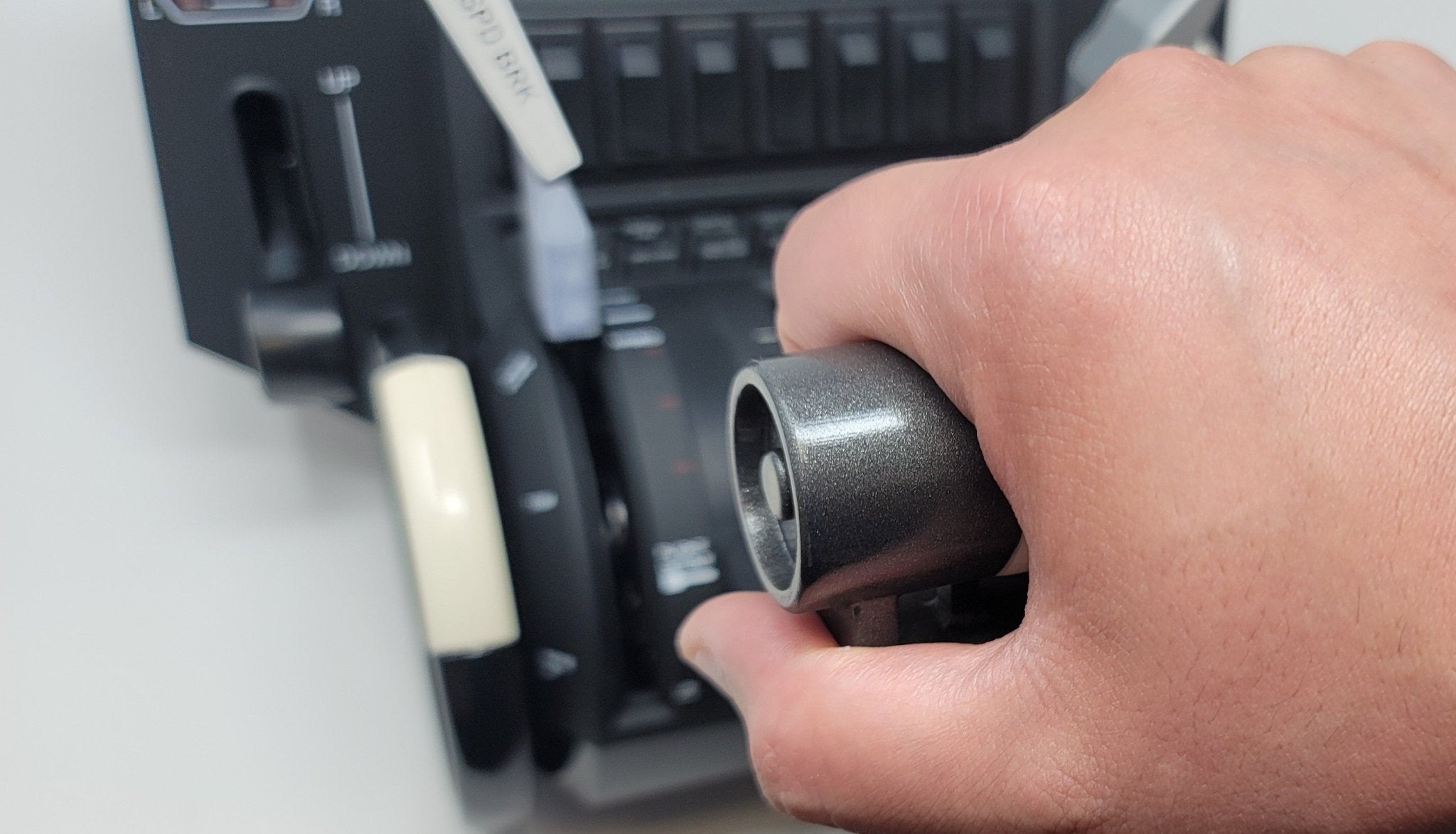 A person is gripping the throttle lever of an aircraft's control panel, reminiscent of a Prodesksim Boeing 757/767 base throttle quadrant addon for the Honeycomb Bravo. The panel features various switches and indicators. The hand firmly holds the black lever, with part of the flight simulation panel visible in the background.