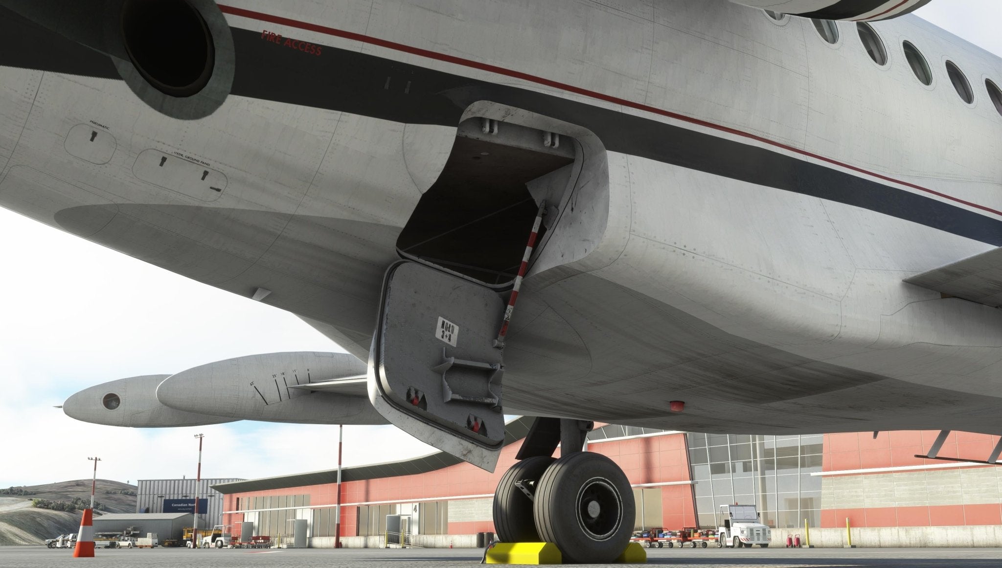 Close-up of a Fokker-28 regional jet’s landing gear and open cargo door at an airport. The aircraft, part of the Just Flight Fokker-28 Professional for MSFS2020 series, is parked near a terminal with a red and gray facade. A bright yellow wheel chock is placed in front of the tire.